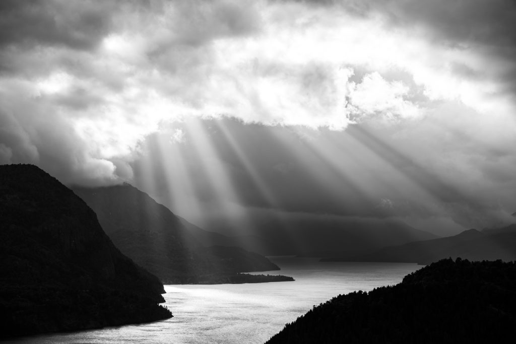 Rayos de sol que atraviesan las nubes sobre un lago y montañas, en blanco y negro